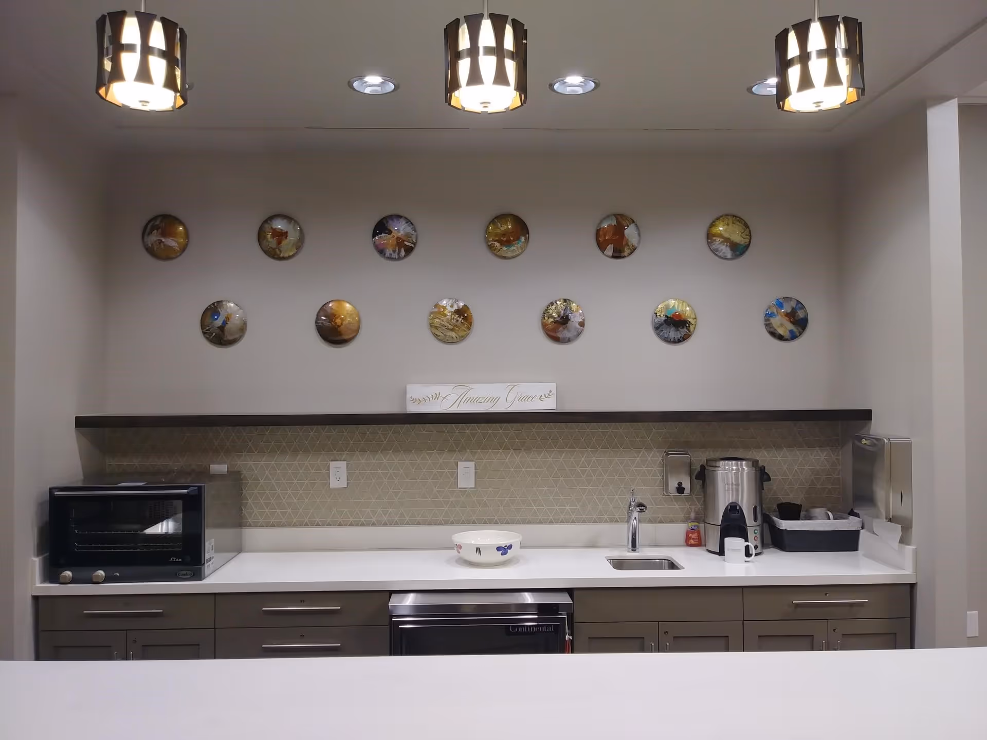 A modern kitchenette area with a white countertop, gray cabinets, a small sink, a coffee urn, a toaster oven, and a bowl. Above the counter, there is a decorative shelf with a sign that reads 'Amazing Grace' and a wall decorated with twelve round artistic plates. Three pendant lights hang from the ceiling.