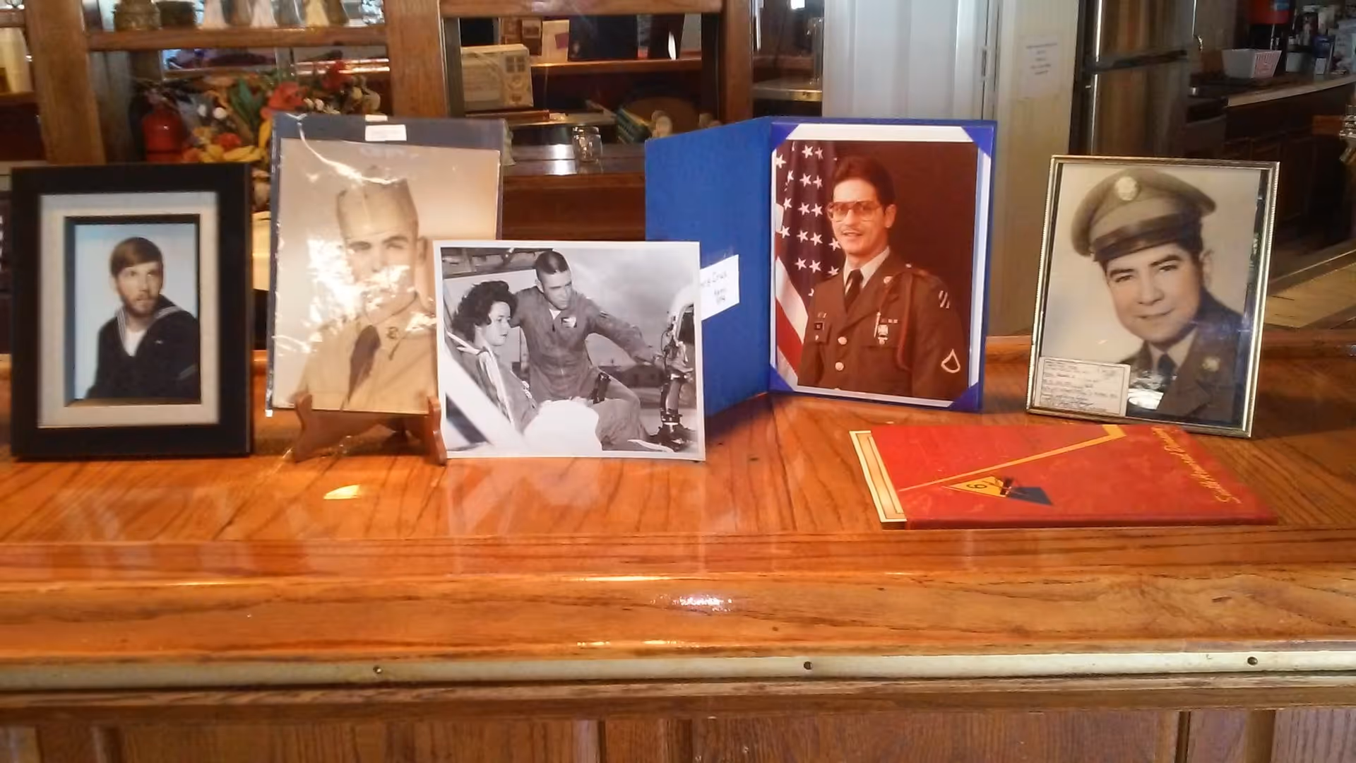 A wooden table displaying framed photographs of men in military uniforms and a black and white photo of a man and woman, along with a red folder or booklet.