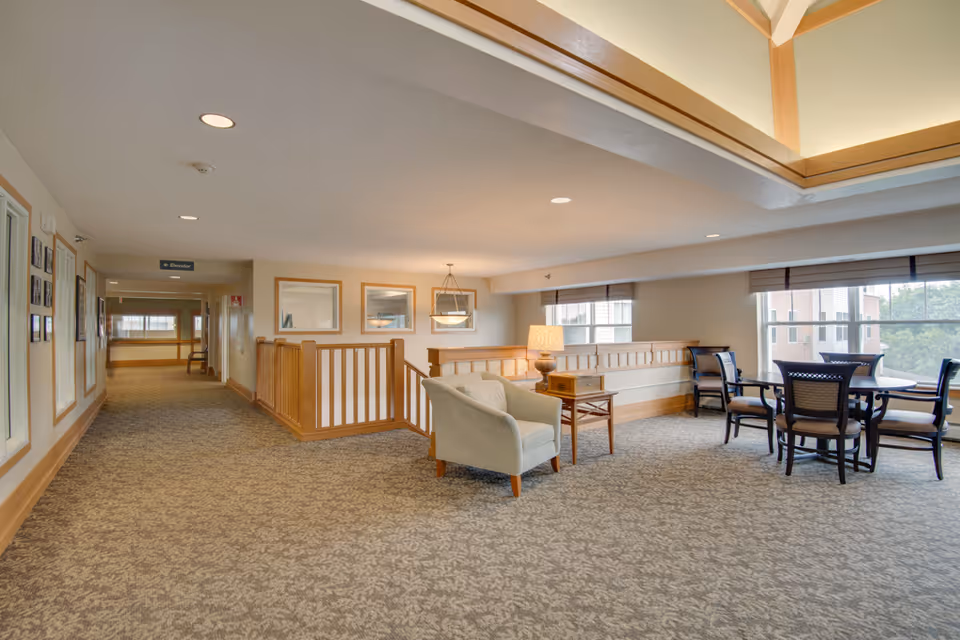 A spacious, well-lit common area in a senior living facility with carpeted floors, beige walls, and wooden trim. The area features a comfortable armchair next to a small wooden table with a lamp, a round table with four chairs near large windows, and a hallway leading to an elevator. The ceiling has recessed lighting and a skylight.