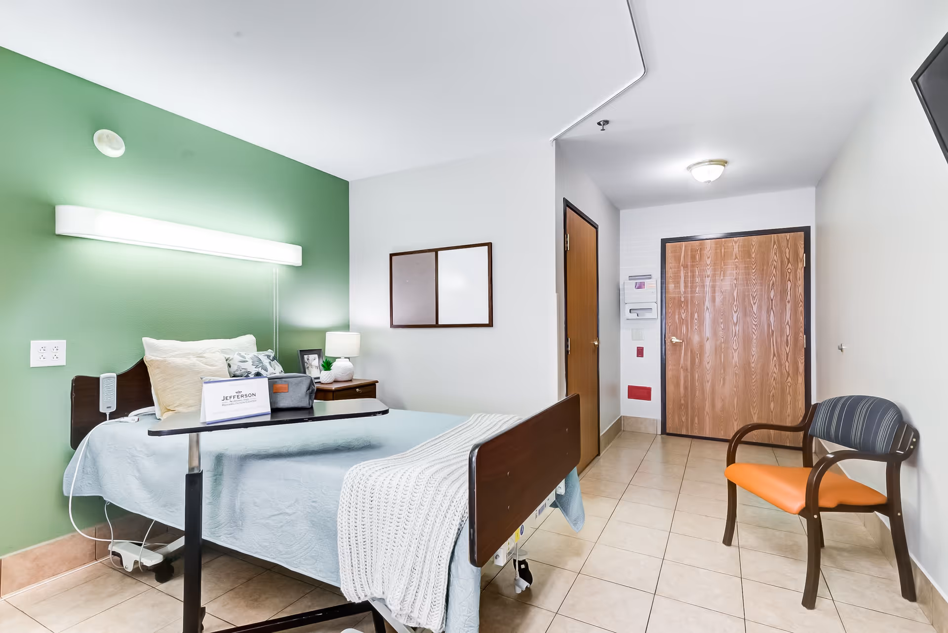 A bright nursing facility patient room with a single bed against a green accent wall, an overbed table, bedside lamp and a chair near the entrance door.