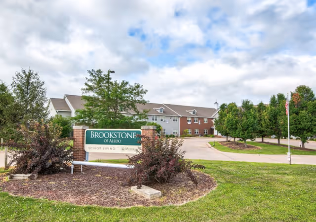 Exterior view of Brookstone of Aledo senior living facility with a large sign in front surrounded by landscaping, a driveway leading to the building, and trees lining the property under a partly cloudy sky.