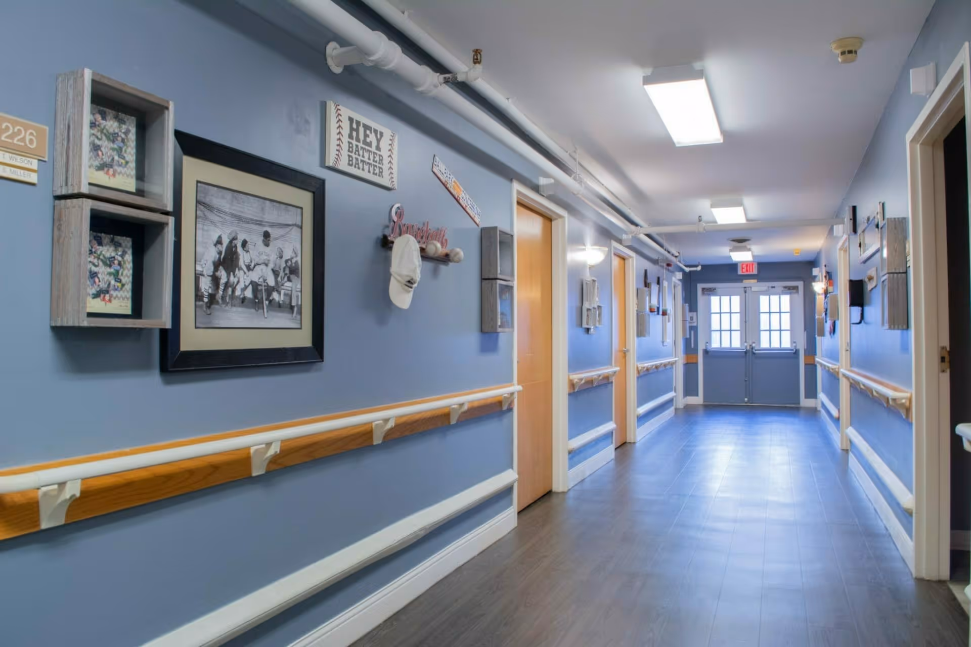 Well-lit blue-painted interior hallway with handrails, framed pictures and doors to resident rooms.