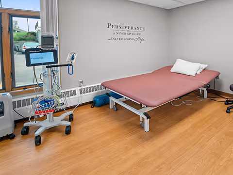 A therapy/treatment room with an examination table, medical monitoring equipment on a rolling stand, and a motivational wall decal.
