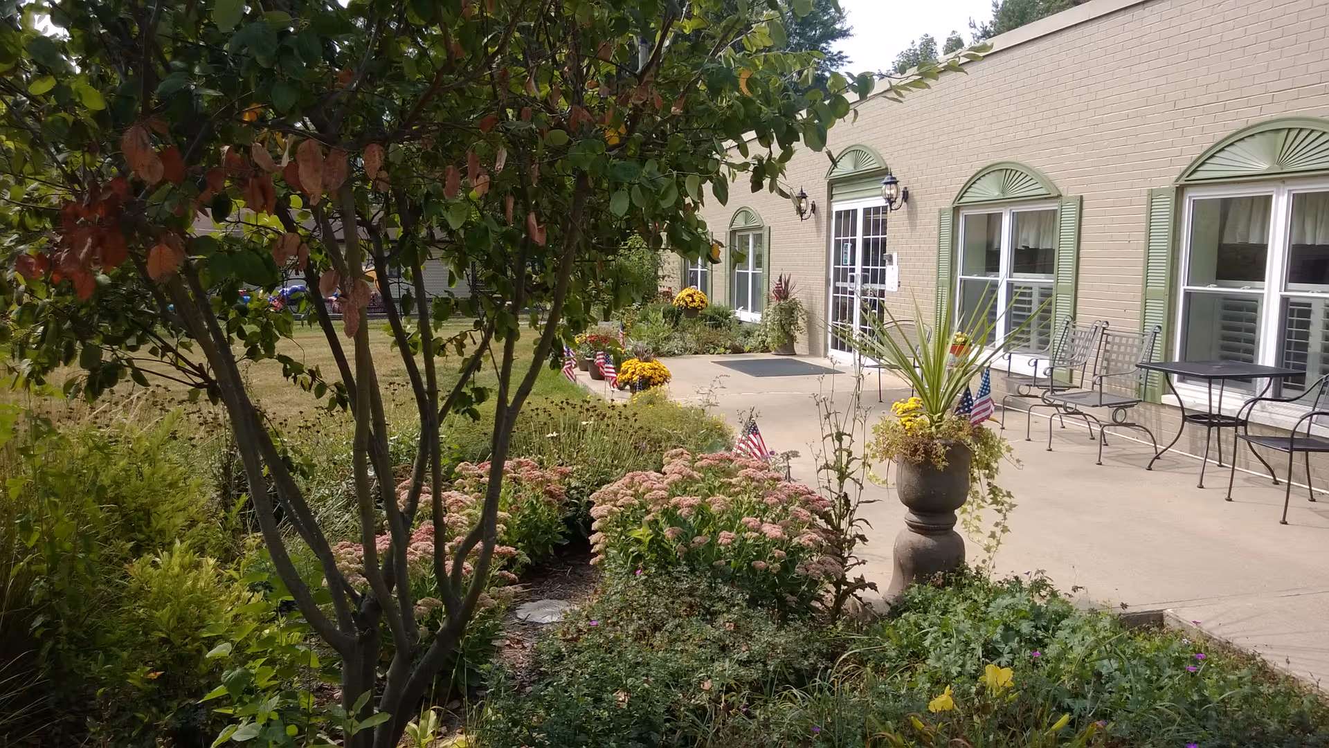 Outdoor garden area with various plants and flowers, including a tree with green and brown leaves in the foreground. A concrete patio with metal chairs and tables is adjacent to a beige brick building with green window shutters and a glass door entrance. Small American flags are placed among the flowers.