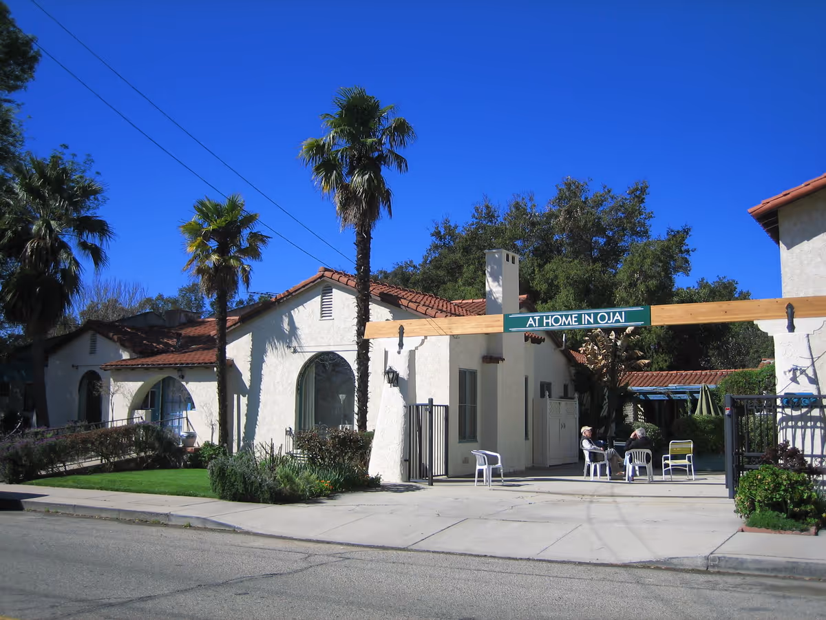 Exterior view of a senior living facility with white stucco walls and red tile roof, palm trees, and a wooden sign that reads 'AT HOME IN OJAI'. Two elderly people are sitting on white plastic chairs in a shaded patio area.