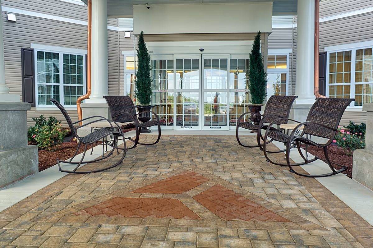 Covered front entrance of a senior living facility with several wicker rocking chairs on a patterned brick porch facing glass double doors.