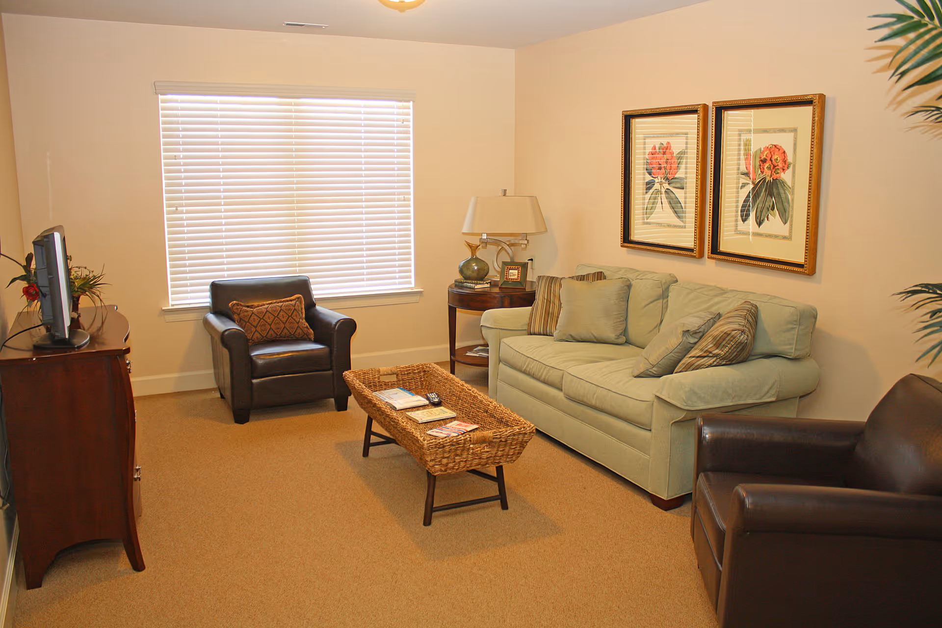 A cozy living room with a light green sofa adorned with several pillows, two dark brown leather armchairs, a wicker coffee table with magazines and a remote control, a wooden TV stand with a television, a side table with a lamp and decorative items, two framed floral prints on the wall, and a window with closed blinds.