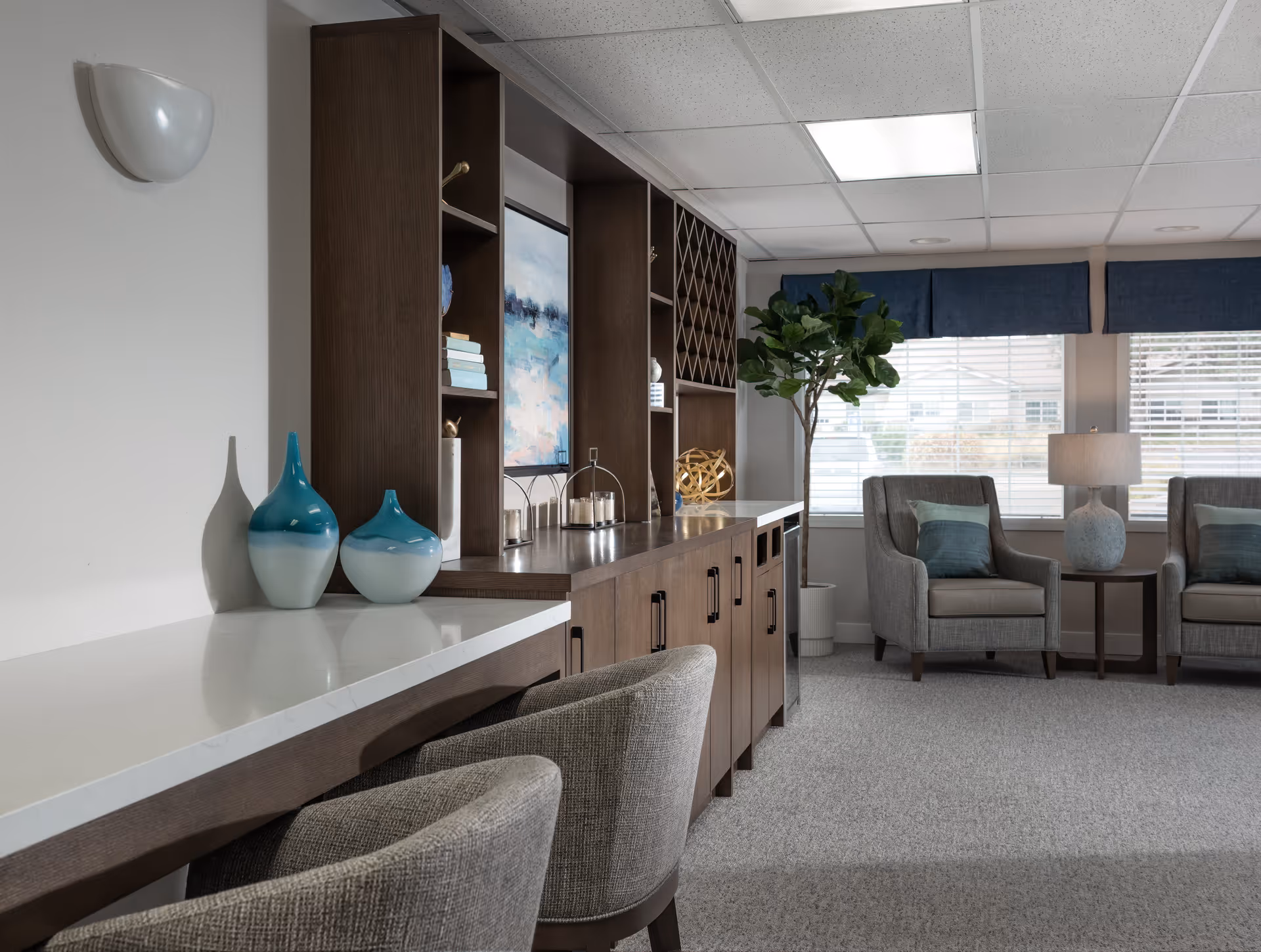 A modern and cozy interior lounge area featuring a long wooden cabinet with shelves and decorative items, two upholstered chairs with cushions, a small side table with a lamp, large windows with blinds and blue valances, and a potted plant in the corner.