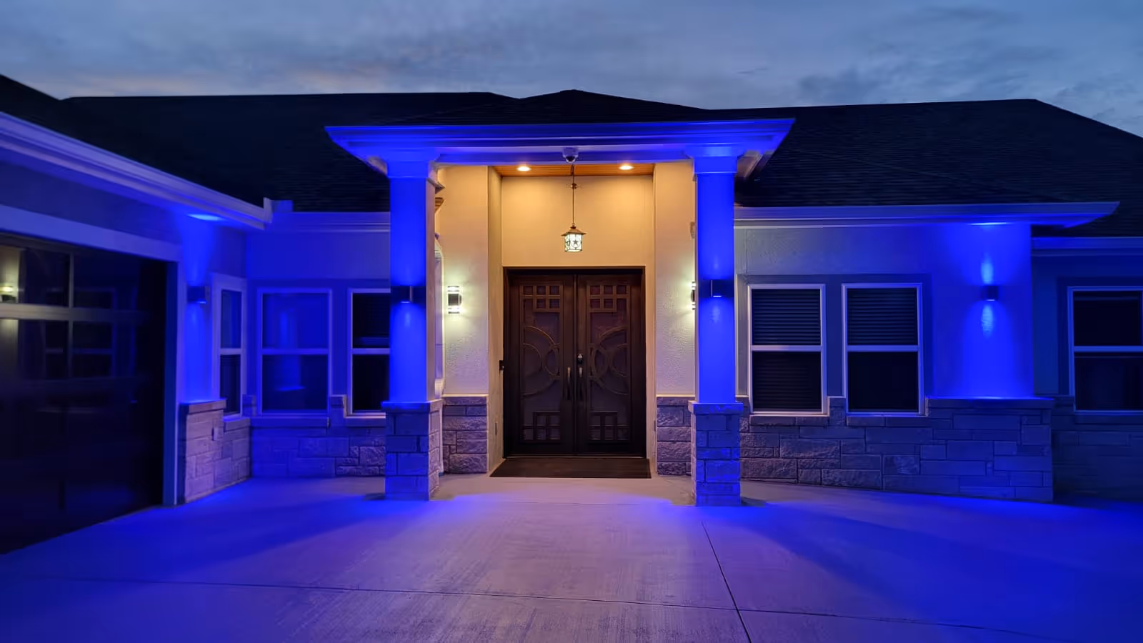 Front entrance of a building at dusk with blue exterior lighting illuminating the walls and columns. The entrance features double wooden doors with decorative glass panels, flanked by stone accents and lit by wall sconces and a hanging lantern above the doors.