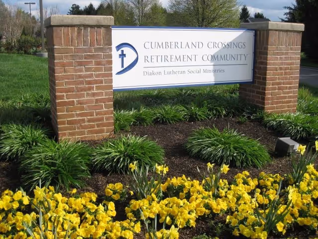 A large sign for Cumberland Crossings Retirement Community mounted between two brick pillars, surrounded by green shrubs and yellow flowers, with trees and a road in the background.