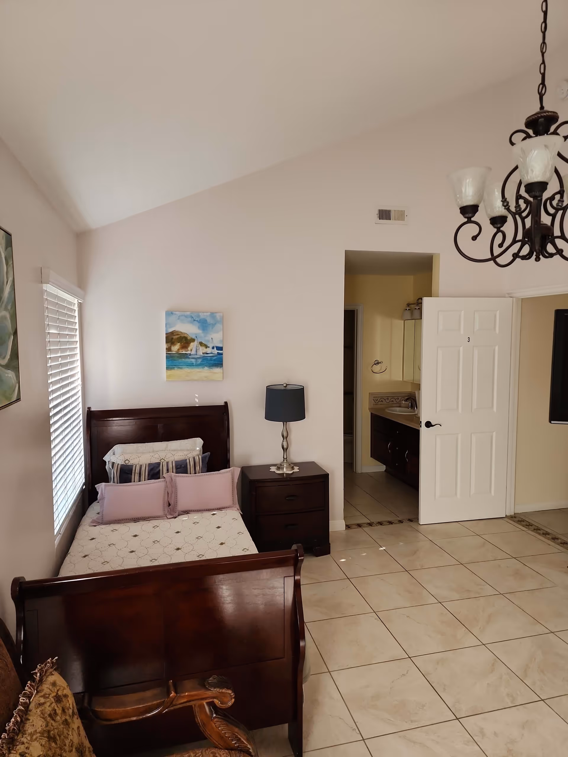 A bedroom with a wooden bed frame and a bed made with white and purple pillows. Next to the bed is a dark wooden nightstand with a lamp. There is a window with blinds on the left wall and a painting of a beach scene above the bed. The floor is tiled with light-colored tiles. An open door leads to a bathroom with a sink and mirror visible. A chandelier hangs from the ceiling.