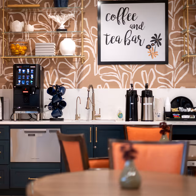 A coffee and tea bar area with a coffee machine, blue mugs on a rotating holder, two airpots for hot beverages, a sink with a modern faucet, and various supplies including paper cups and condiments. The wall behind features a decorative beige and white pattern with a framed sign that reads 'coffee and tea bar'. In the foreground, there are orange cushioned chairs and a round table with a small vase holding a flower.