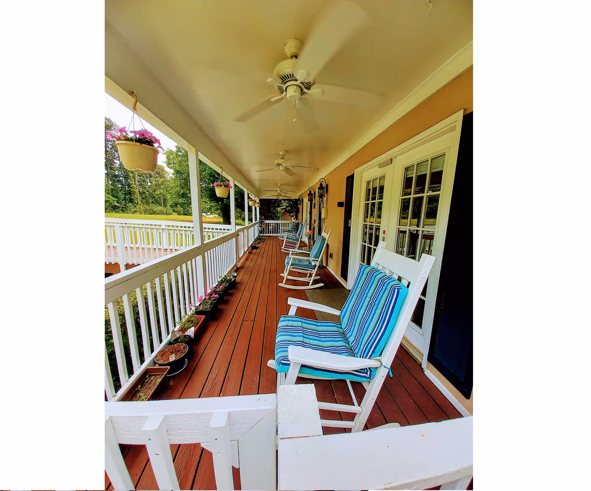 Long covered wooden porch with white railing, multiple rocking chairs with blue-striped cushions, hanging flower pots, and ceiling fans.