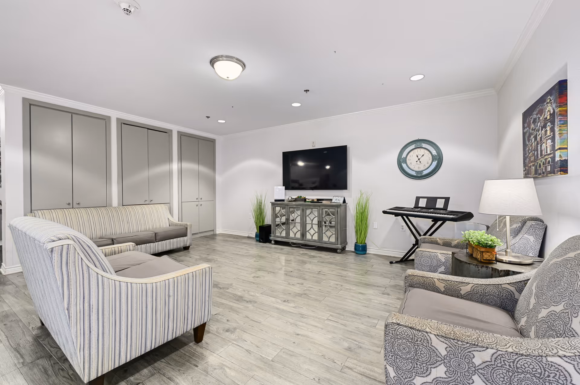 A bright and spacious living room with light gray wood flooring, two striped sofas, two patterned armchairs, a round side table with a lamp and plant, a wall-mounted flat screen TV above a mirrored cabinet, a keyboard on a stand, a round wall clock, and a colorful painting on the wall.