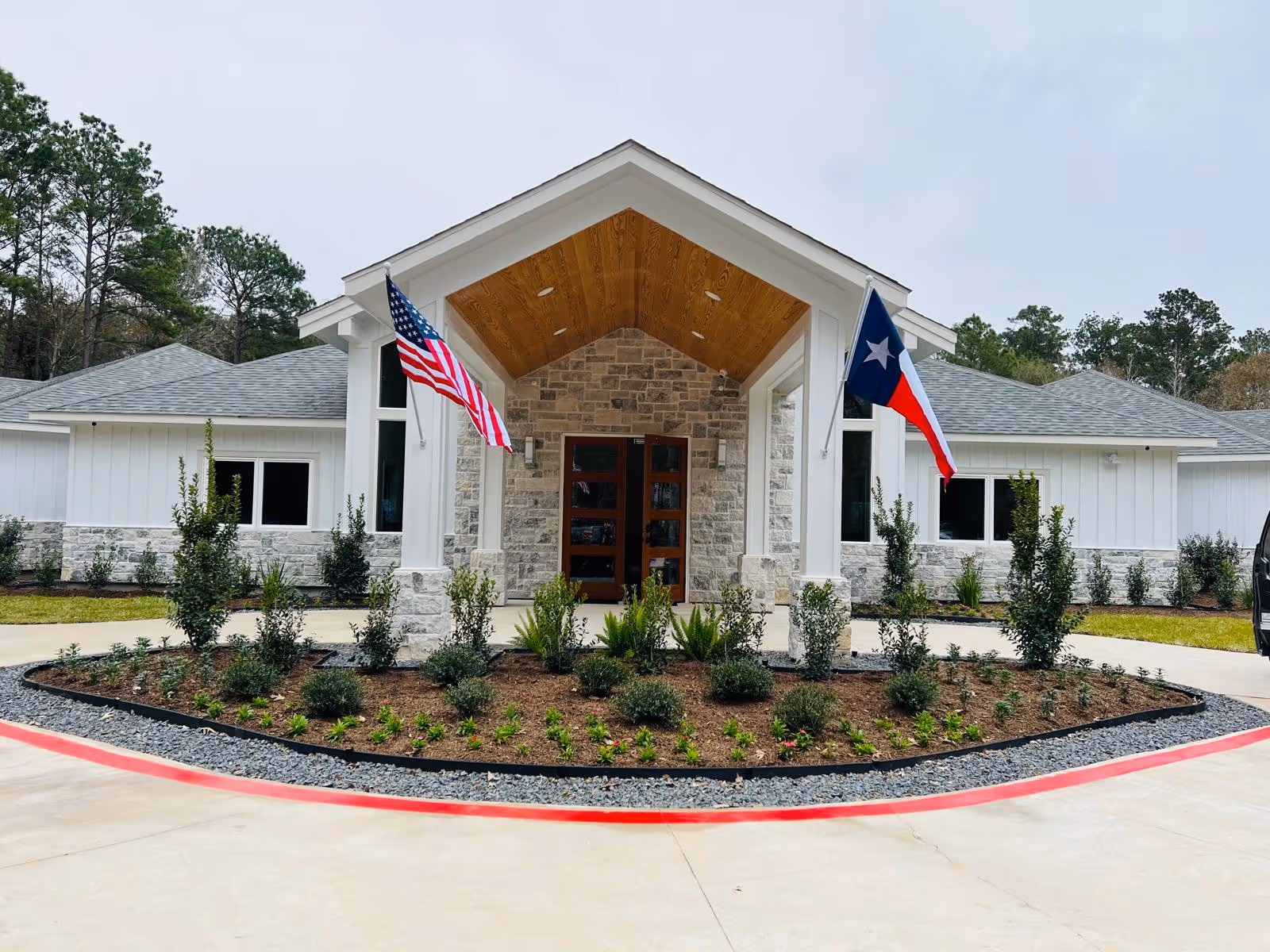 Front entrance of a single-story assisted living building with a covered portico, landscaped circular bed, and American and Texas flags.