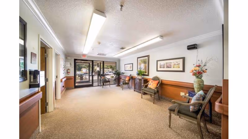 A well-lit interior common area with beige carpet and white walls featuring framed artwork. There are several chairs with cushions, a small table with a plant, and a large vase with flowers. The far end has glass doors leading outside, and there is a fire extinguisher mounted on the wall near the exit.