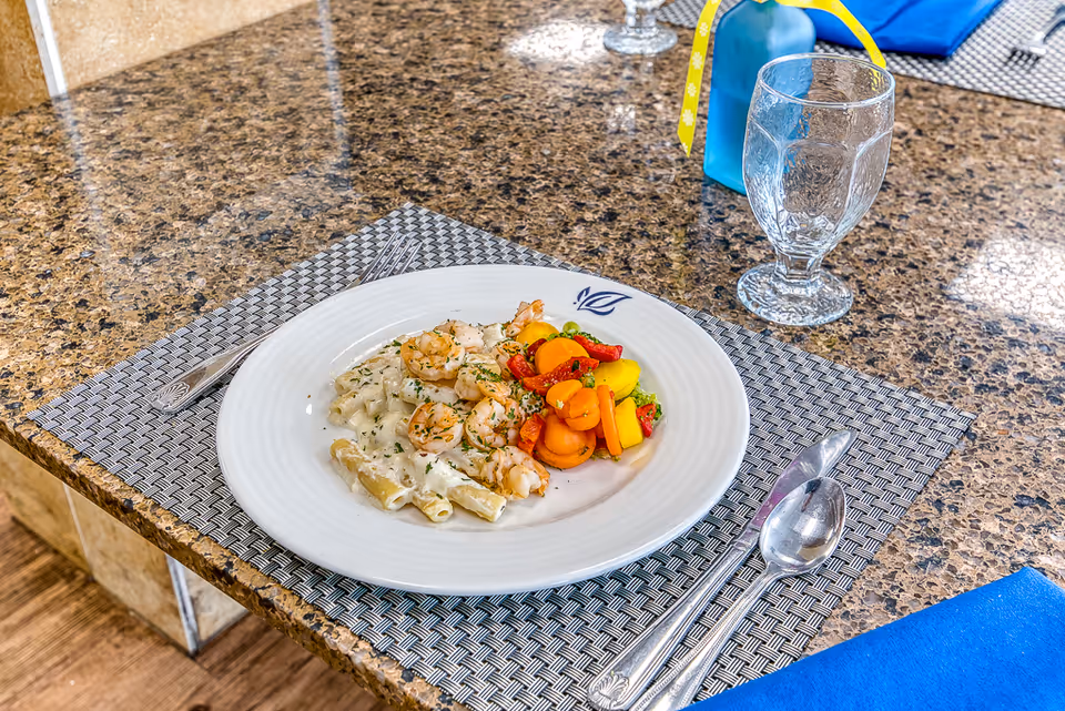 A plate of creamy pasta with shrimp and a side of mixed cooked vegetables on a woven placemat on a granite countertop. There is a fork and spoon on either side of the plate, a clear glass, and a blue napkin with a small blue bottle in the background.