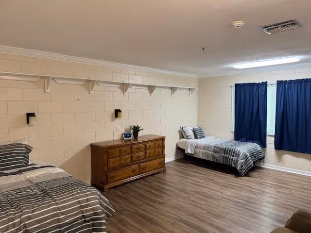 A simple bedroom with two single beds on opposite sides of the room, each with striped bedding. Between the beds is a wooden dresser with a small plant and a framed photo on top. The walls are light-colored brick, and there is a long shelf with hooks above the dresser. The floor is wood laminate, and a window with dark blue curtains is on the far wall.