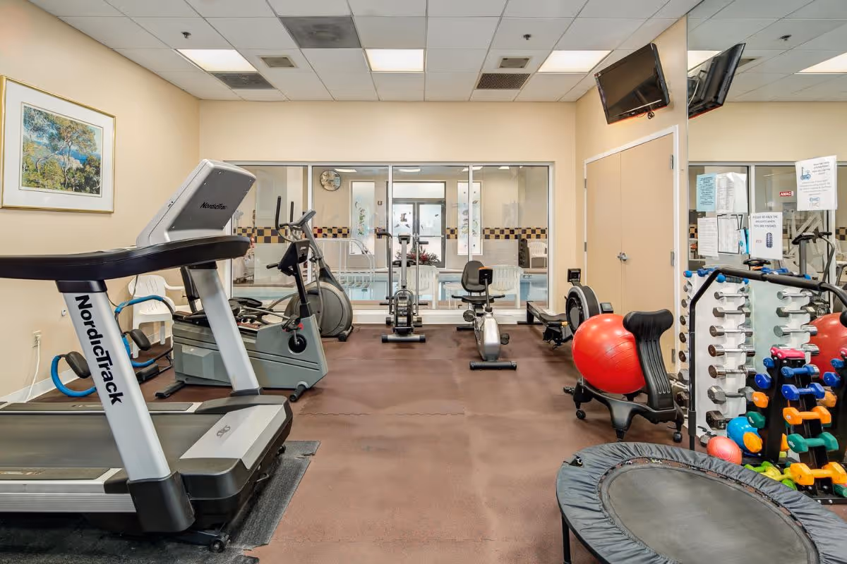 Indoor fitness room with exercise equipment including a treadmill, stationary bikes, rowing machine, weights, exercise balls, and a small trampoline. A large window shows an indoor pool area in the background. The room has beige walls, a wall-mounted TV, and a large mirror reflecting the equipment.