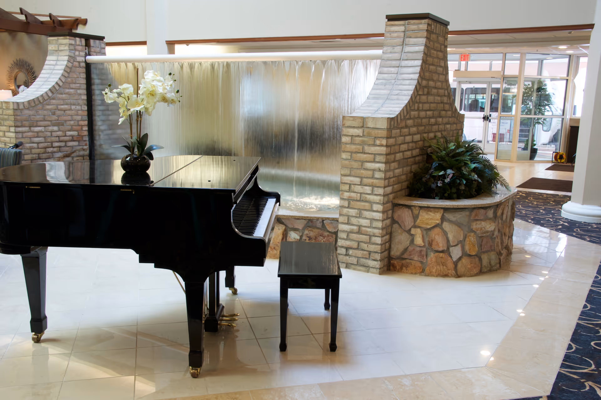 A black grand piano with a matching bench is placed on a polished tile floor in a spacious indoor area. Behind the piano, there is a decorative water feature with water cascading down a glass wall. To the right, a brick and stone planter holds green plants. The background shows glass doors leading outside and part of a carpeted area with a blue and beige pattern.