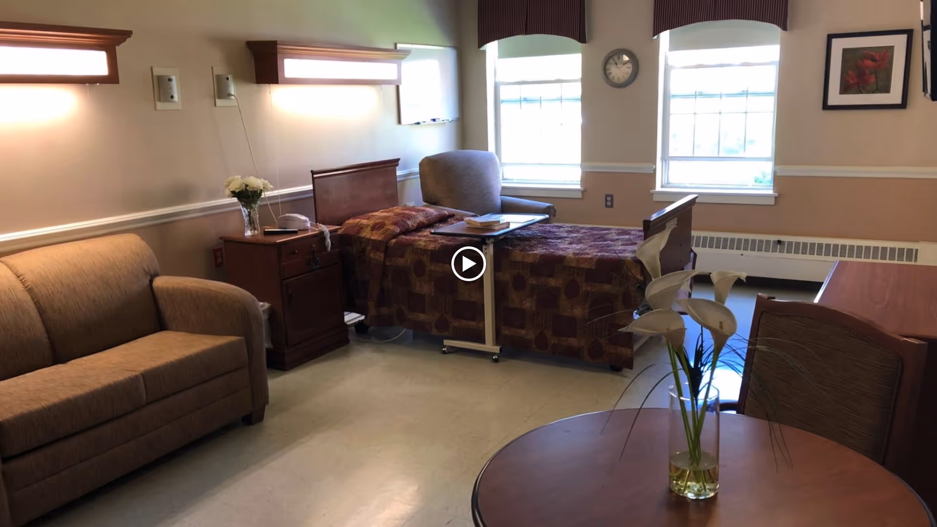 A well-lit room with a single bed covered in a patterned quilt, a bedside table with a telephone and flowers, a comfortable armchair, a sofa, and a round table with a vase of white flowers. Two windows with valances let in natural light, and a clock and framed artwork hang on the wall.