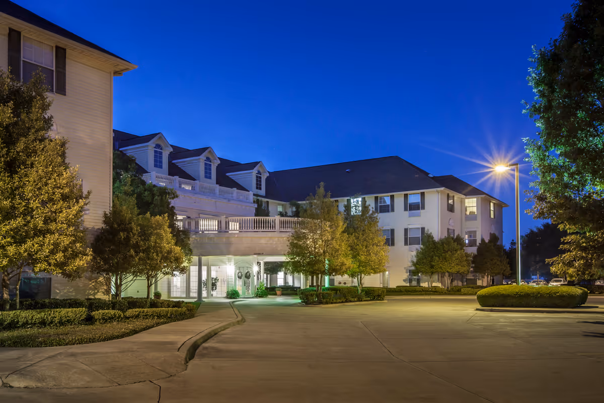 Exterior view of a senior living facility named Bloom at Bossier at dusk, showing a well-lit building with multiple windows, trees, and a driveway with street lamps.