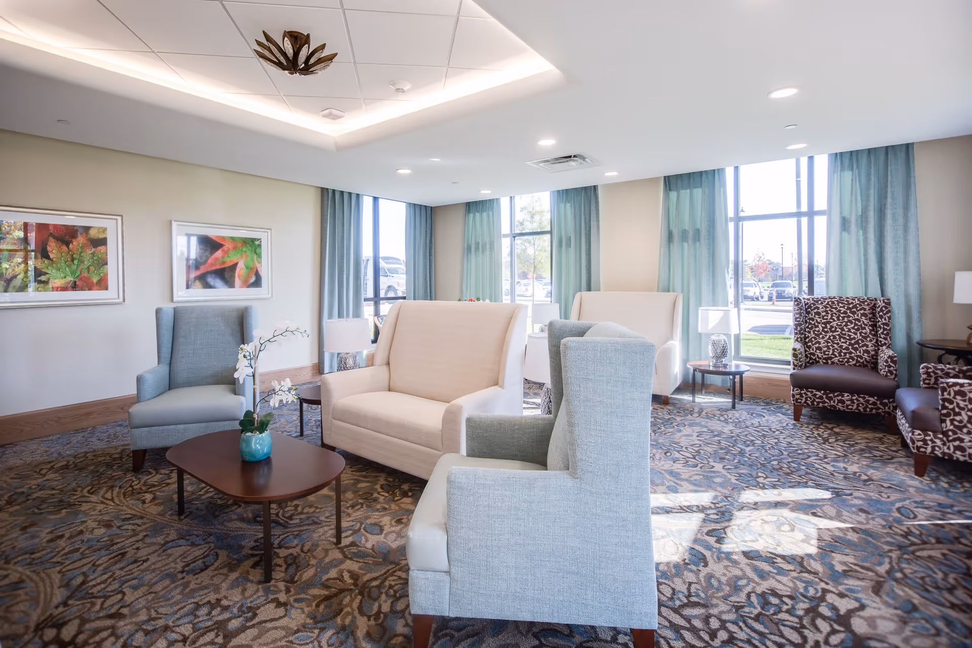 A bright and spacious living room area with multiple upholstered chairs and loveseats arranged around a wooden coffee table with a small potted orchid. Large windows with blue curtains allow natural light to fill the room. The walls are decorated with framed artwork featuring colorful leaves, and the ceiling has recessed lighting and a decorative light fixture.