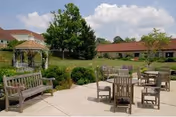 Outdoor patio area with wooden benches and tables with chairs on a concrete surface, surrounded by green bushes and trees, with buildings and a gazebo in the background under a partly cloudy sky.