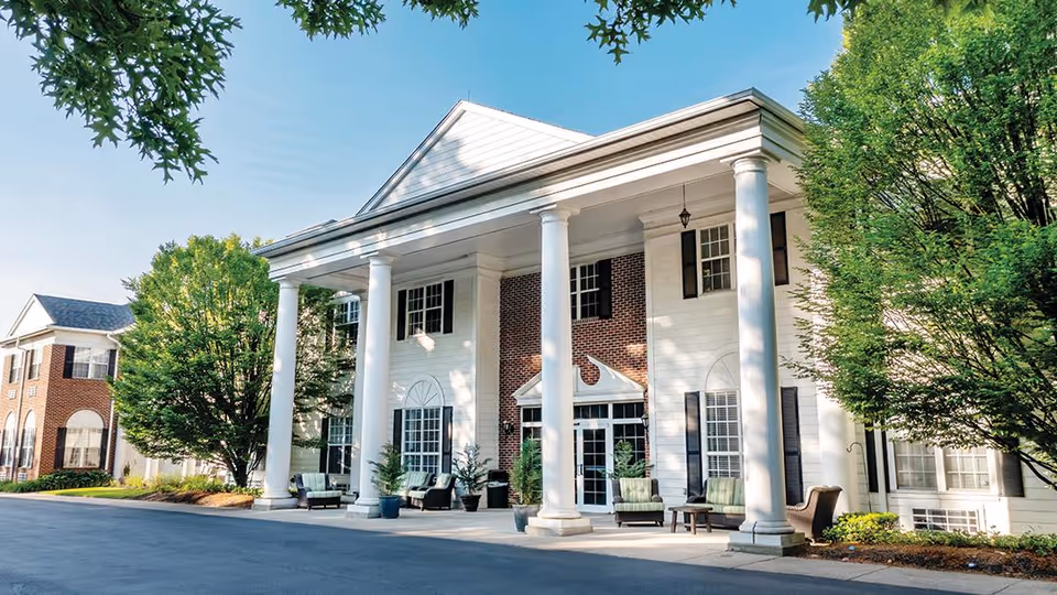 Exterior view of a senior living facility named Blue Bell Place featuring a large white building with tall white columns, brick accents, multiple windows with black shutters, outdoor seating, and surrounded by green trees and landscaping.