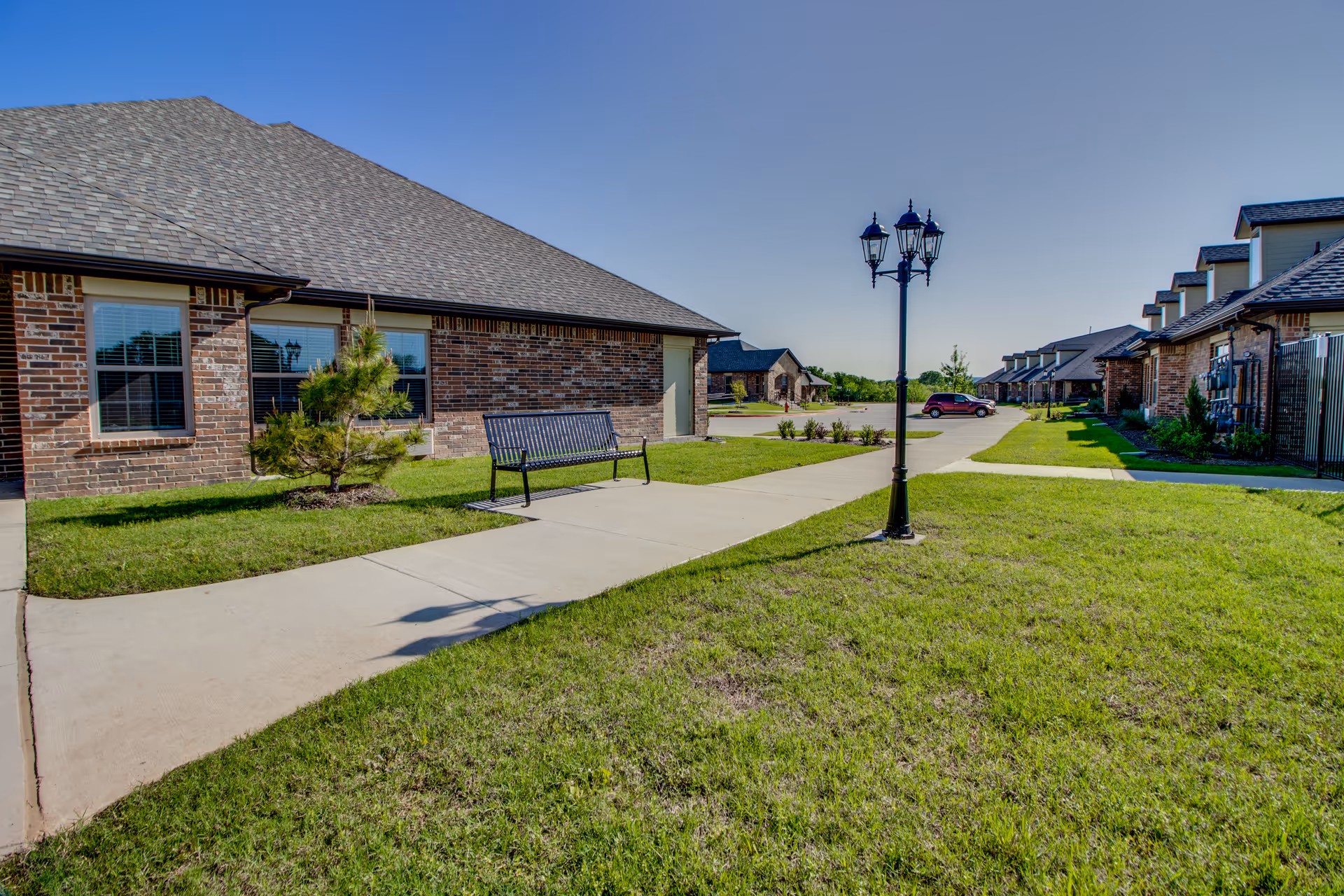 Brick single-story assisted living buildings flank a paved walkway with a bench, lamp post, and green lawns under a clear sky.