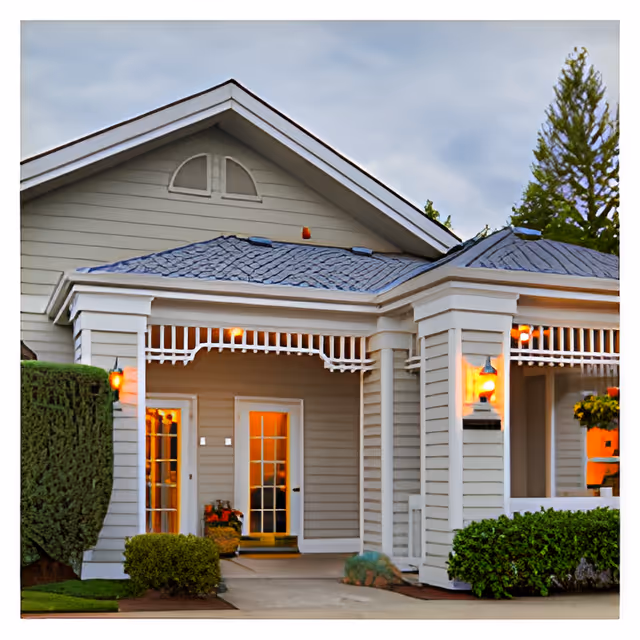 Exterior view of a senior living facility building with beige siding, white trim, and a gray shingled roof. The entrance features a covered porch with decorative white railings and warm lights glowing near the doors and windows. There are neatly trimmed bushes and a tree visible around the building.
