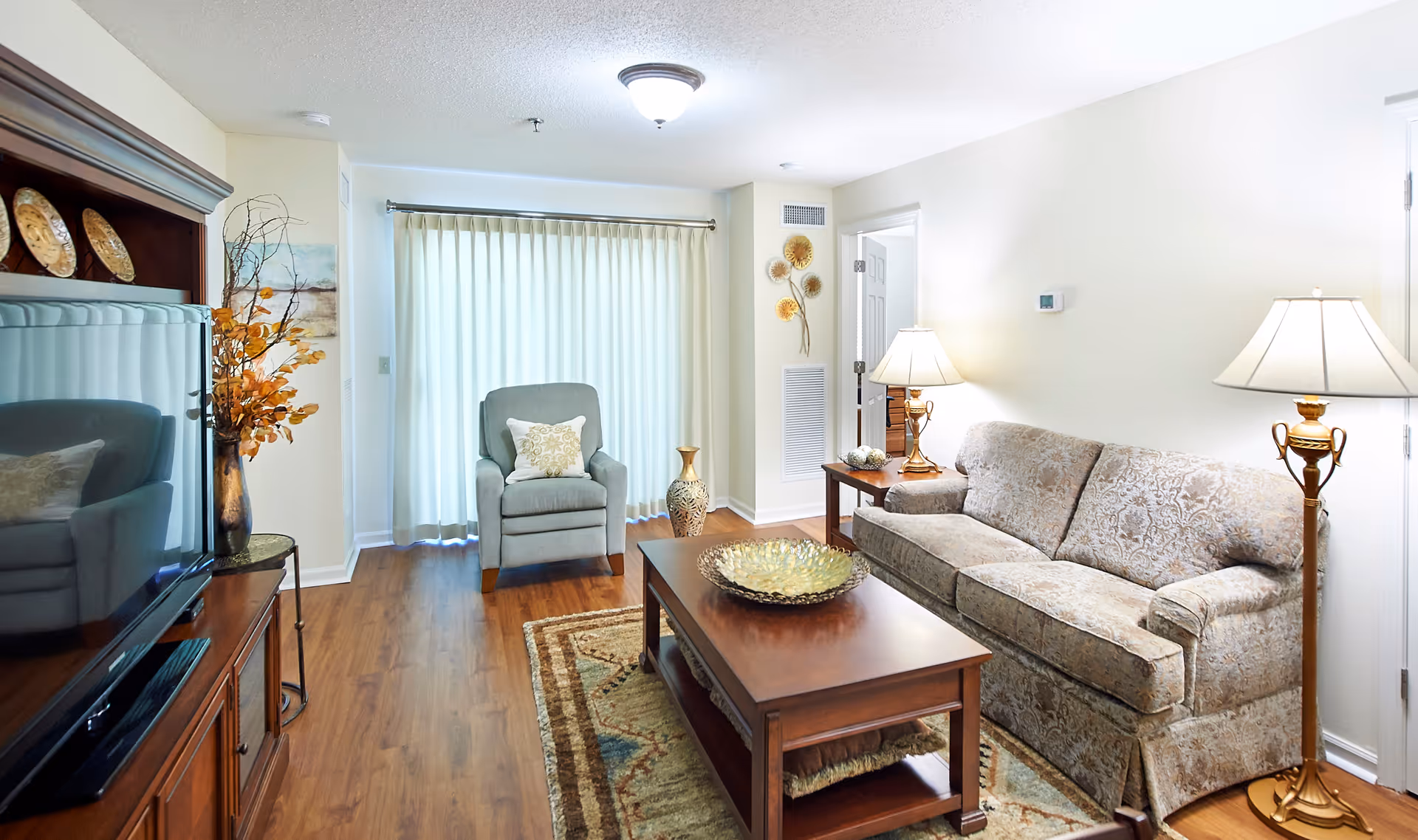 A bright and cozy living room with a patterned beige sofa, a wooden coffee table with a decorative bowl, a gray armchair with a cushion, two table lamps, a floor lamp, a large TV on a wooden stand, and a window covered with sheer curtains. The room has wooden flooring and light-colored walls with minimal decor.