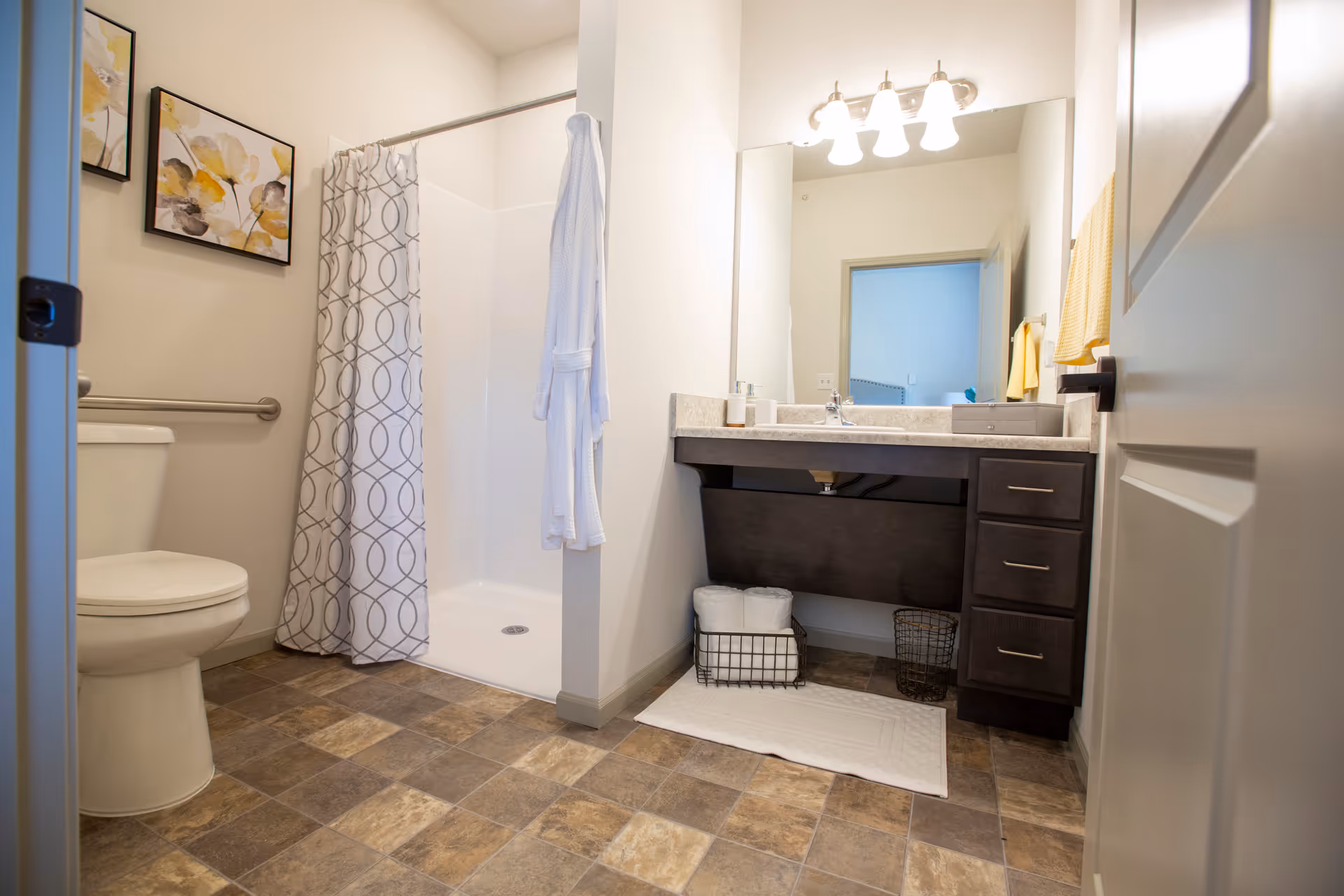 A clean and modern bathroom featuring a toilet, a walk-in shower with a patterned curtain, a vanity with a large mirror, three drawers, and a countertop with a soap dispenser. There are two framed floral artworks on the wall above the toilet, a white bathrobe hanging on the shower curtain rod, and a white bath mat on the floor in front of the vanity. The floor has a tile pattern in shades of brown and beige.