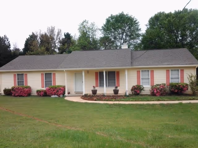 Single-story beige house with a gray roof, white front door, and red window shutters. The front yard has green grass, a curved concrete walkway, and several bushes with pink flowers. Trees are visible in the background.
