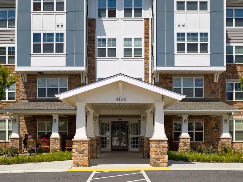 Front exterior view of a multi-story senior living facility with a covered entrance supported by stone pillars. The building features large windows and a combination of stone and siding on the facade. The address number 4120 is displayed above the entrance.