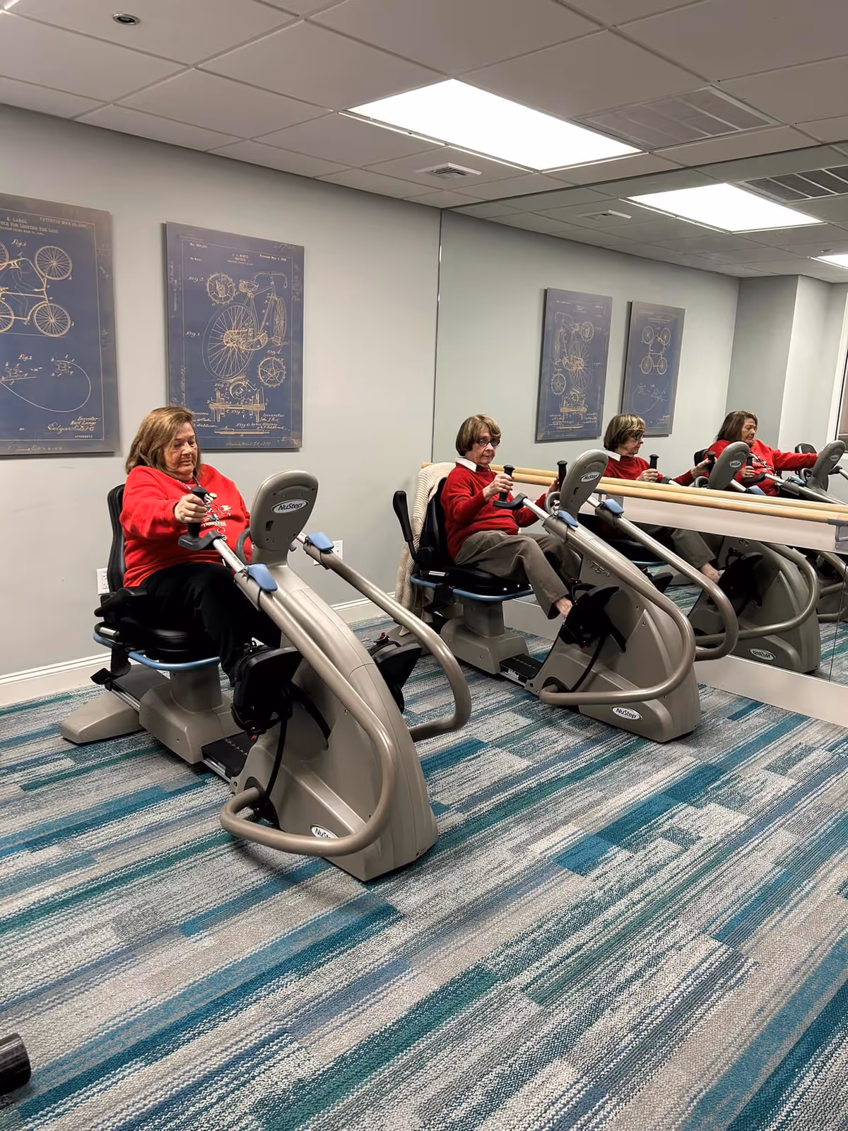 Two elderly women in red sweaters exercising on seated elliptical machines in a fitness room with a large mirror on the wall and framed bicycle blueprint artwork.