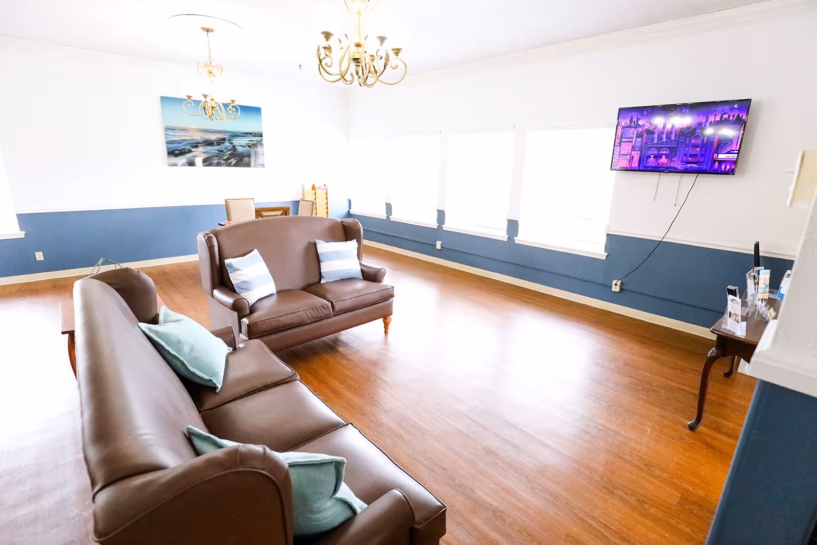 A bright living room with wooden floors, two brown leather sofas with light blue and white pillows, a wall-mounted flat screen TV, a chandelier, and a large window letting in natural light. There is a small table with brochures on the right side and a framed picture of a beach scene on the wall.