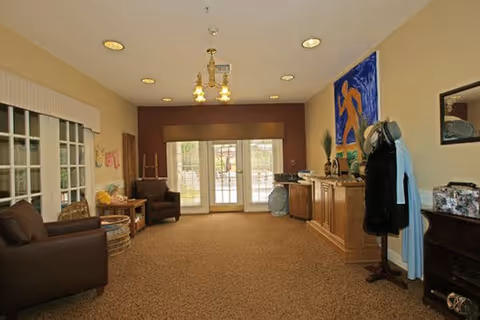 Interior room with beige walls and carpeted floor featuring two brown armchairs on the left, a wooden cabinet with decorative items on the right, a coat rack with clothing, a large colorful painting on the wall, and double glass doors leading outside at the far end.