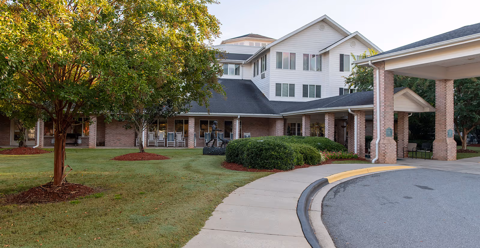 Exterior view of a multi-story senior living building with a covered entrance, landscaped lawn, and trees.
