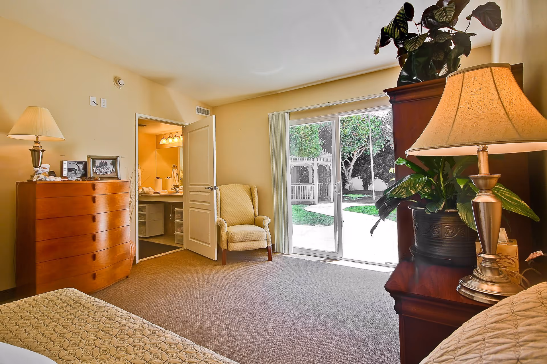 A cozy bedroom in a senior living facility with a wooden dresser topped with a lamp and framed photos, a comfortable armchair near an open door leading to a bathroom, and a sliding glass door opening to a garden with a white gazebo and trees.