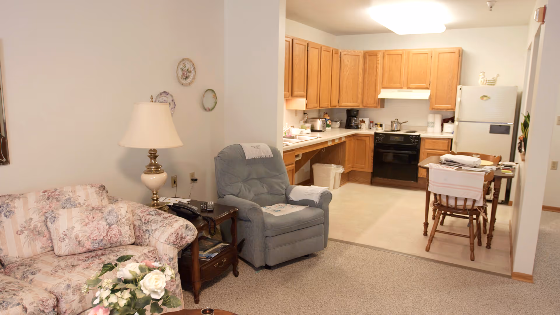 A cozy living area with a floral patterned sofa, a side table with a lamp and telephone, and a blue recliner chair. Adjacent to the living area is a kitchen with wooden cabinets, a black stove, a white refrigerator, and a small dining table with two chairs. The walls are light-colored and decorated with small plates.