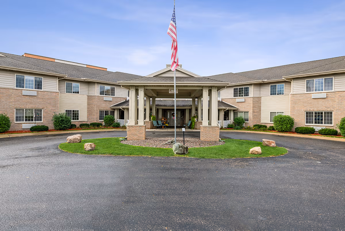 Front exterior view of a two-story senior living facility with a covered entrance supported by columns. An American flag is flying on a flagpole in front of the entrance. The building has a combination of brick and siding with multiple windows and landscaping including bushes and rocks around the driveway.