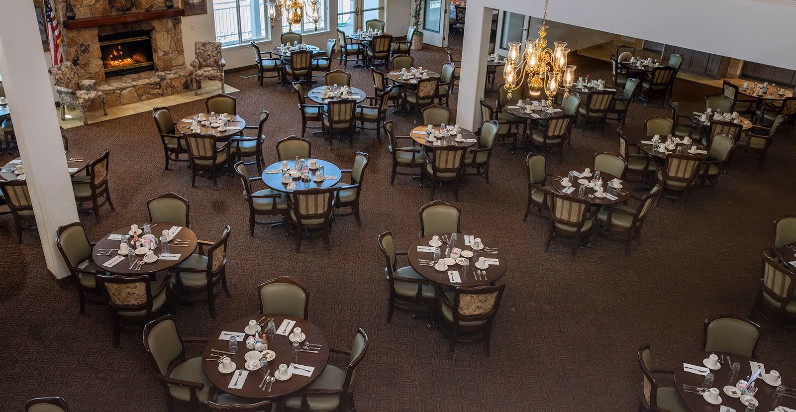 A spacious dining room with multiple round tables set with cups, plates, utensils, and napkins. The room features upholstered chairs, a stone fireplace with a fire burning, large windows, and a chandelier hanging from the ceiling.