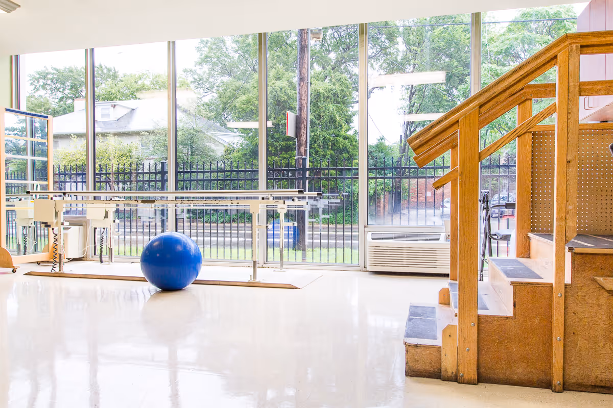 A bright rehabilitation room with large floor-to-ceiling windows showing an outdoor view. Inside, there is a blue exercise ball on the floor, parallel bars for walking support, and a wooden staircase with handrails for physical therapy exercises.