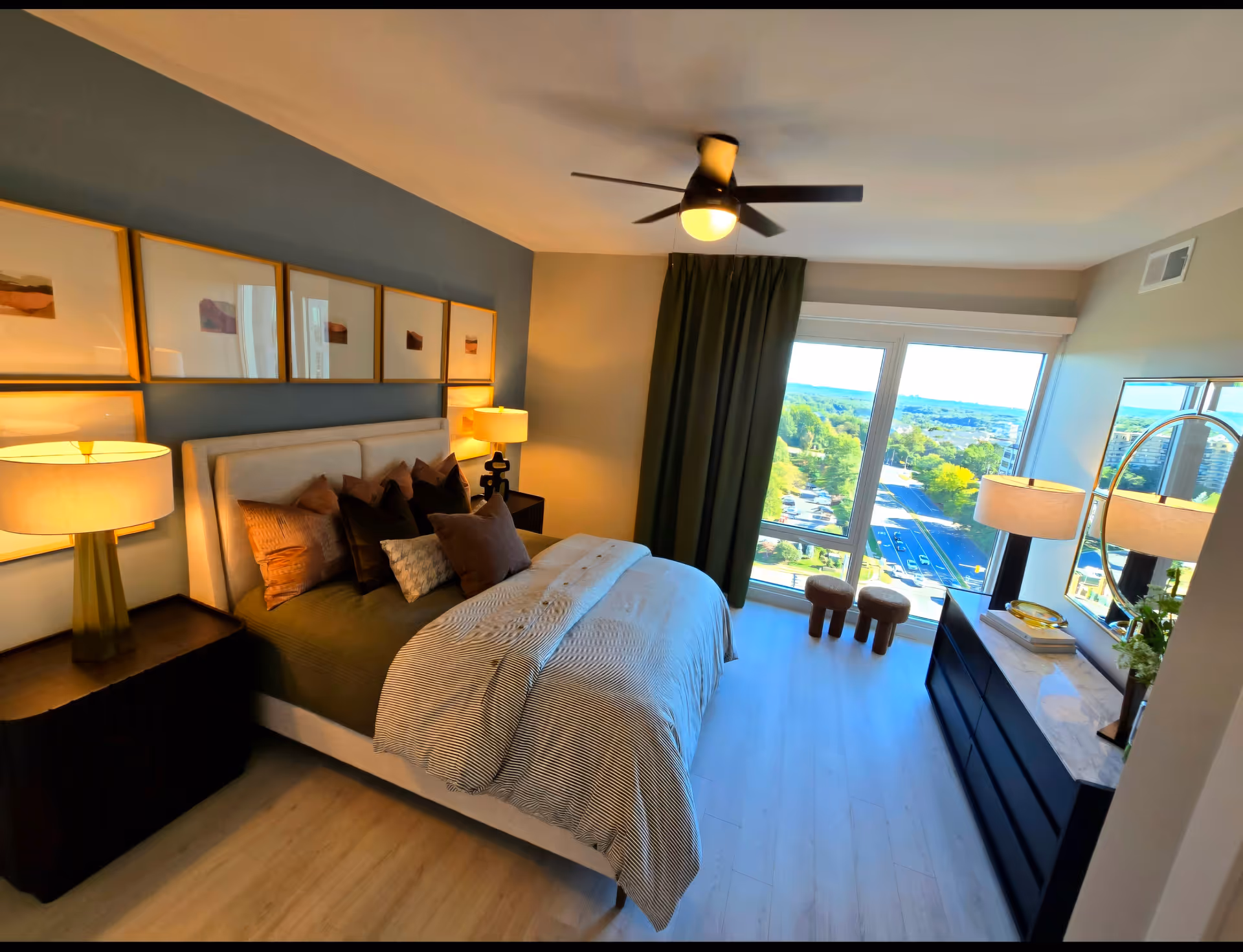 Modern furnished bedroom with a large bed, bedside lamps, dresser and a floor-to-ceiling window overlooking a leafy view.
