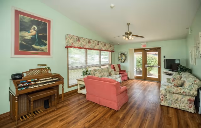 A bright common living room with floral and pink upholstered sofas and armchairs, a wooden organ, TV, ceiling fan, and glass doors to the outside.