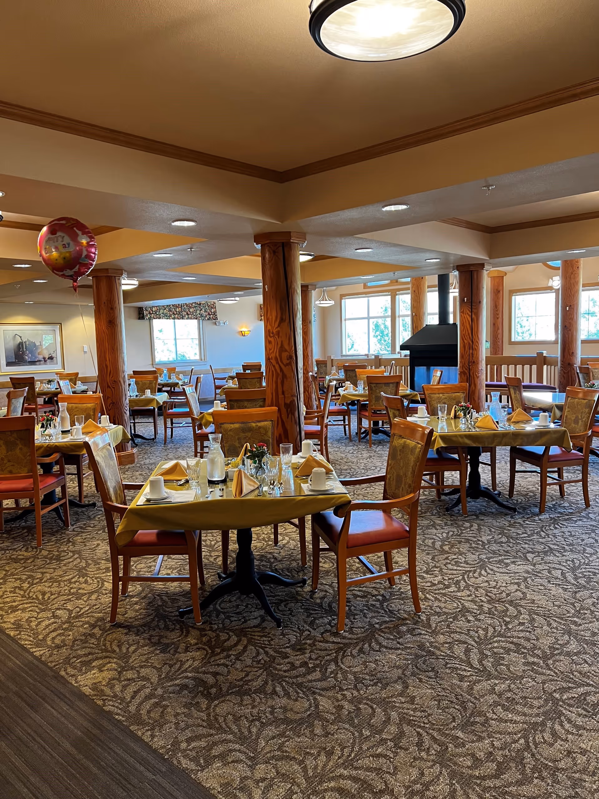 A dining room in a senior living facility with multiple tables set with yellow tablecloths, napkins, glasses, and cups. The room features wooden chairs with cushioned seats, large windows allowing natural light, wooden pillars, and a carpeted floor with a floral pattern. A pink balloon is floating near the ceiling on the left side of the image.