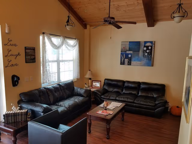 Cozy communal living room with two black leather sofas, a coffee table, side tables, wall art, and a wooden vaulted ceiling with a ceiling fan.