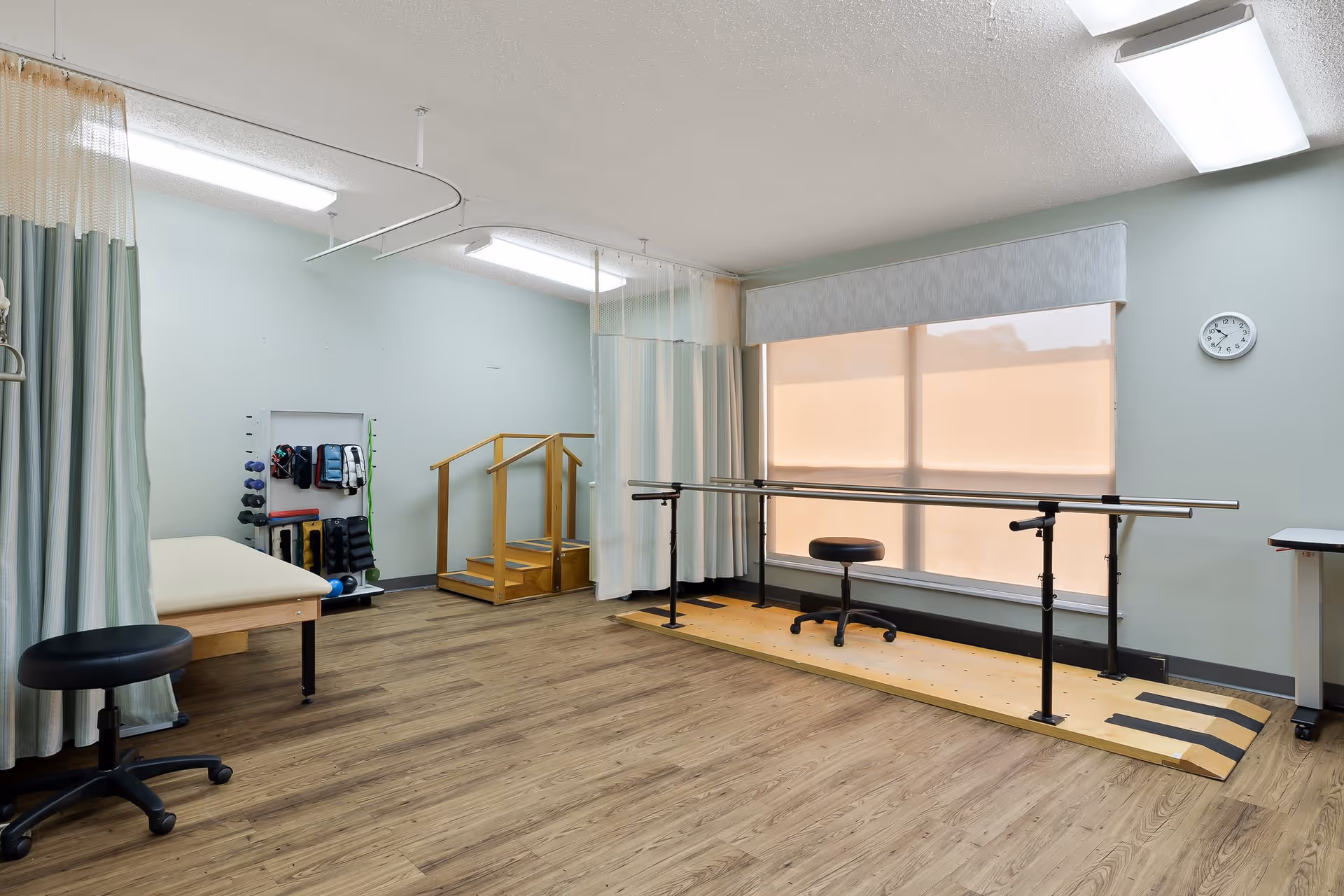 A physical therapy room with wooden flooring, parallel bars for walking exercises, a small set of stairs with handrails, an examination table, stools, and exercise equipment organized on a rack. The room has light blue walls, large windows with blinds, and ceiling-mounted fluorescent lights.