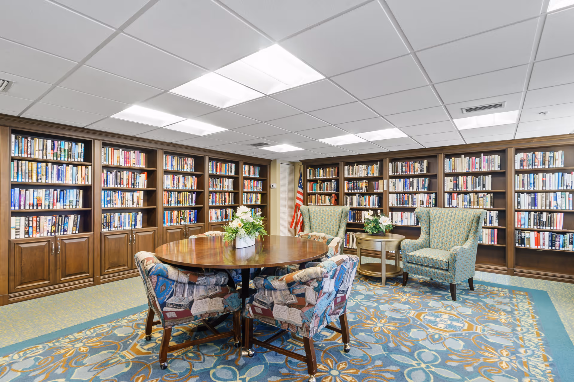 A well-lit library room with wooden bookshelves filled with books lining the walls. In the center, there is a round wooden table with a flower arrangement and four upholstered chairs with a colorful pattern. Two green upholstered armchairs and a small round wooden side table with a plant are also present. An American flag is visible in the corner.